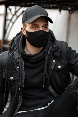 Şık, yakışıklı, tıbbi maskeli ve siyah kapüşonlu siyah ceketli genç adam sokakta oturuyor. Havalı şehir erkek tarzı ve covid-19 konsepti