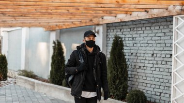 Şık, yakışıklı, koruyucu tıbbi maskeli, şık siyah ceketli, kapüşonlu ve şapkalı genç bir adam şehirde geziyor.