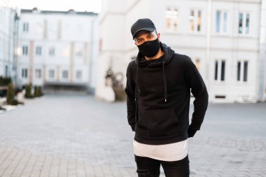 Koruyucu maskeli, siyah şapkalı, şık siyah kapüşonlu yakışıklı bir adam. Kentsel erkek giyim tarzı ve salgın