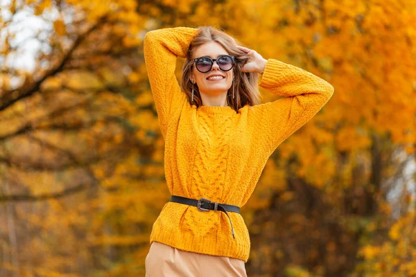 鮮やかなオレンジの葉を持つ森の中でスタイリッシュなサングラスの散歩と流行のニット黄色のセーターで笑顔で幸せな美しい若い女性 — ストック写真