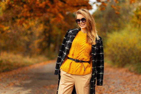 穿着时尚外套 头戴墨镜 头戴黄色复古毛衣 笑容满面的快乐小女孩 走在秋天的公园里 绿叶斑斓 — 图库照片
