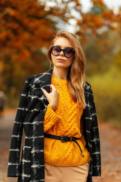 エレガントなファッション美しい女性でスタイリッシュなサングラスでヴィンテージ黄色のニットセーターと黒のコートはカラフルな黄金の葉と秋の自然の中で歩く — ストック写真