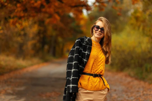 身穿时髦的黄色针织毛衣 头戴外套 戴着太阳镜 满意地笑着来到一个有着橙色秋天树叶的公园 — 图库照片