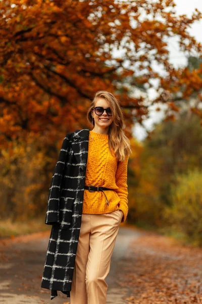 ファッションコートのかわいい女性の顔と黄色のニットセーターのきれいな笑顔の女性モデルは 紅葉と秋の公園で歩きます 幸せな感情 — ストック写真