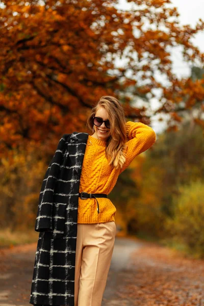 身穿黄色古色古香毛衣 黑色外套和太阳镜的快乐微笑的女孩 穿着时髦的秋装 带着五彩缤纷的落叶走进了公园 — 图库照片