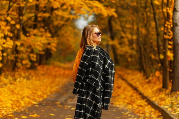漂亮的欧洲女人 戴着时髦的太阳镜 穿着时髦的黑色外套 在一个迷人的公园里 有着明亮的黄色秋叶 — 图库照片