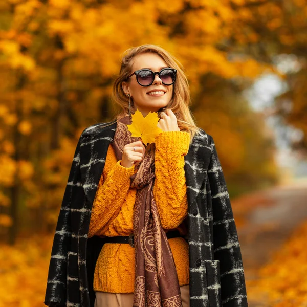 美丽的年轻女子 穿着时髦的秋装 身穿黑色外套 头戴针织毛衣 头戴墨镜 笑容可亲 在黄色的落叶公园里挂着一片秋叶 — 图库照片