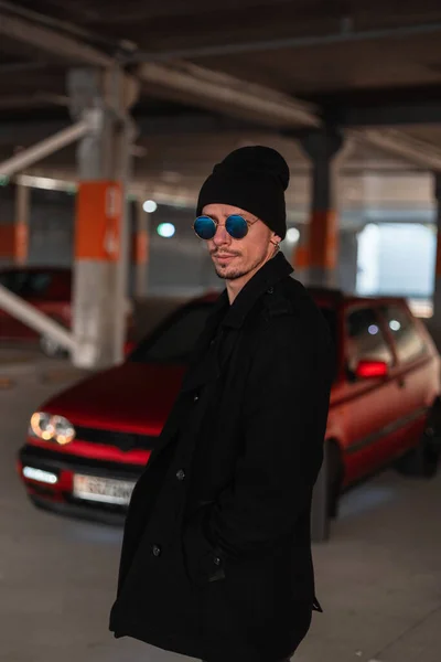 若いですファッション男でスタイリッシュな青サングラスでヴィンテージコートと帽子近くに古い赤車 — ストック写真