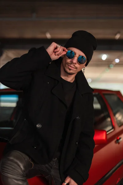都会の赤い車の近くに帽子をかぶったエレガントな黒のコートにクールなサングラスを持つファッショナブルな若い男 — ストック写真