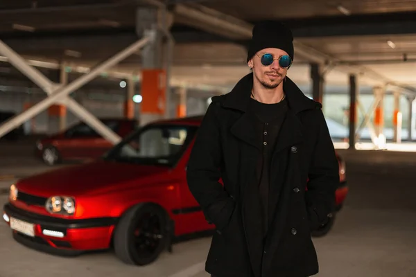 ファッションサングラスと駐車場で市内の赤い車の近くのヴィンテージブラックコートの帽子を持つスタイリッシュなハンサムな若い男モデル — ストック写真