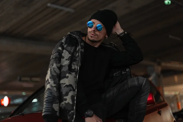 Portrait of a handsome young hipster guy with cool sunglasses in a fashionable winter military jacket with a black pullover and hat stands near a red car in the city