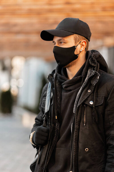 Fashionable young man model with a protective medical mask in stylish clothes, winter jacket with a hoodie and a backpack walks in the city