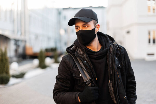 Fashionable handsome hipster man with a black cap and an inter-military protective mask in a black jacket, hoodie with backpack walks in the city. Male modern urban style and pandemic concept