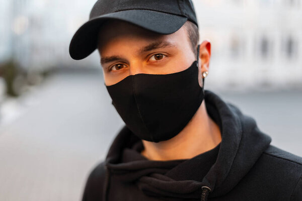 Urban fashion portrait of handsome young guy with protective medical mask and black cap in stylish casual hoodie outdoors. COVID-19
