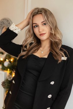 Lüks ceketli şık siyah takım elbiseli zarif, genç bir bayan model beyaz duvarın yanında duruyor.