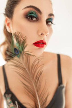 Profesyonel makyajlı, kırmızı dudaklı ve yeşil göz kapaklarıyla beyaz arka planda yüzün yanında renkli bir tavus kuşu tüyü tutan güzel bir kızın yaratıcı portresi. Cilt bakımı ve güzellik