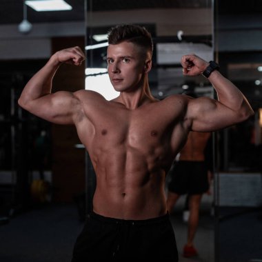 Çıplak kaslı gövdesi olan yakışıklı sporcu vücut geliştirici ve karanlık bir arka planda spor salonunda poz veriyor.