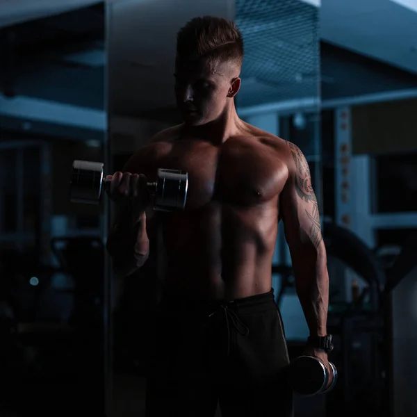 年轻英俊的健身教练 身材矮小 肌肉发达 在黑暗的体育馆里用哑铃做运动 — 图库照片