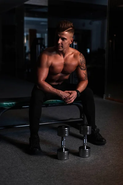 在黑暗中 年轻英俊的运动健将 身材矮小 肌肉发达 坐在体育馆的哑铃旁 运动锻炼 — 图库照片