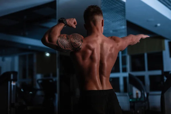 身强体壮的健美运动员 身材矮小 肌肉发达 在黑暗中背对着体育馆里的摄像机站着 — 图库照片