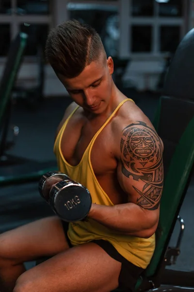 年轻的健美健美运动员 带着玛雅纹身 坐在体育馆里给他的二头肌抽气 — 图库照片