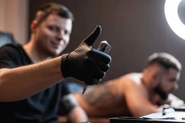 Professional tattoo master man hipster in black gloves with a tattoo machine shows like thumbs up and makes a tattoo