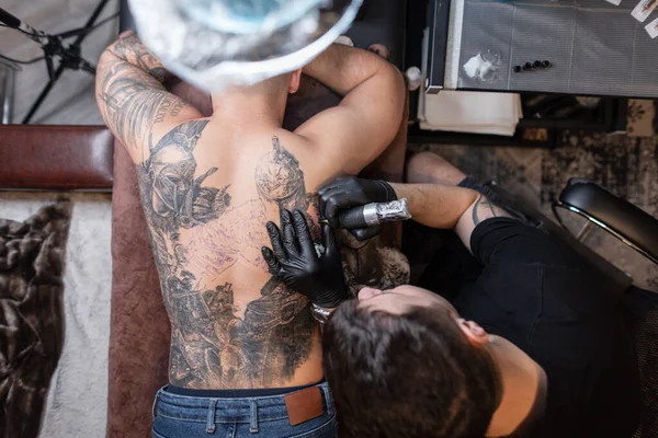 Man professional tattoo artist with black gloves and a modern tattoo machine makes a tattoo with ink on a male back, top view. tattoo creation workflow