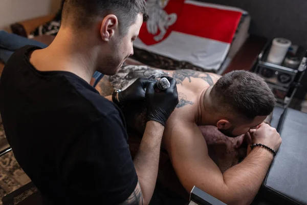 Man professional tattoo artist in black gloves makes a tattoo on the male back in the studio. Creative work process