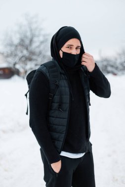 Siyah moda yeleği, kapüşonlu, şapkalı ve siyah moda sırt çantalı siyah koruyucu maskeli yakışıklı erkek model kışın karlı bir günde seyahat ediyor. Coronavirus ve tarz kavramı
