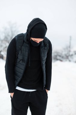 Koruyucu maskeli yakışıklı genç bir adam, şık, siyah bir kışı dışarıda, sırt çantalı, açık havada kar altında yürüyor.