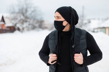 Modern portrede, bir kış günü şehirde, siyah bir elbise, yelek, kapüşonlu, şapka ve sırt çantalı, yakışıklı bir genç adam dolaşır. Coronavirus ve dikkatli seyahat kavramı.