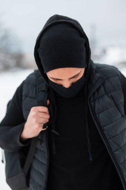 Siyah koruyucu tekstil maskeli şık bir adam. Sırt çantalı siyah elbiseli. Bir kış günü sokakta kar altında yürüyor.
