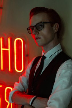 Gözlüklü, şık bir genç adam. Şık bir iş elbisesi var. Üzerinde kırmızı neon yazılar olan yeşil bir duvarın yanında kravatı var.