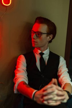 Moda sektöründe gözlüklü, yelek, gömlek ve kravat takan şık bir ofis çalışanı modeli kırmızı ışıklı yeşil bir duvarın yanında oturuyor.