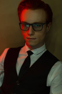 Şık iş elbiseleri, gömleği, yeleği ve kravatıyla gözlüklü şık bir kızıl saçlı güzel bir adamın şık portresi yeşil ve kırmızı ışıkla duvarın yanında oturuyor. Havalı İş Adamı