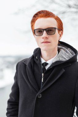 Kırmızı saçlı, gözlüklü, şık iş elbiseli, ceketli, gömlekli ve kravatlı İngiliz genç adam şehirde yürüyor.