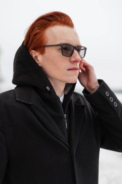 Şık, kızıl saçlı, gözlüklü, ceketli ve kapüşonlu şık bir hippi sokakta beyaz bir arka planda kapüşon takıyor.