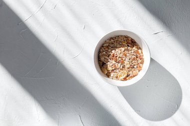 Beyaz kâse, beyaz bir tabakta müsli ile nefis bir kahvaltı, güneş ışığı ve gölgeler, üst manzara. Sağlıklı çiğ vejetaryen yemeği