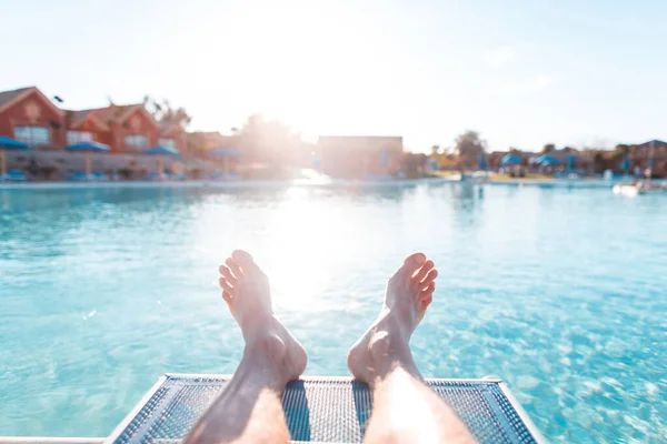 年轻人在日落时躺在游泳池边的海滩上休息 人的脚踏在蓝色的水和宾馆平房的背景上 第一人称观点放松 — 图库照片