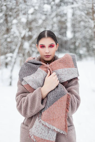 穿着时髦冬衣 头戴围巾 五颜六色 富有创造性地化妆 眼影粉红的年轻貌美女子在雪地森林里摆姿势 — 图库照片