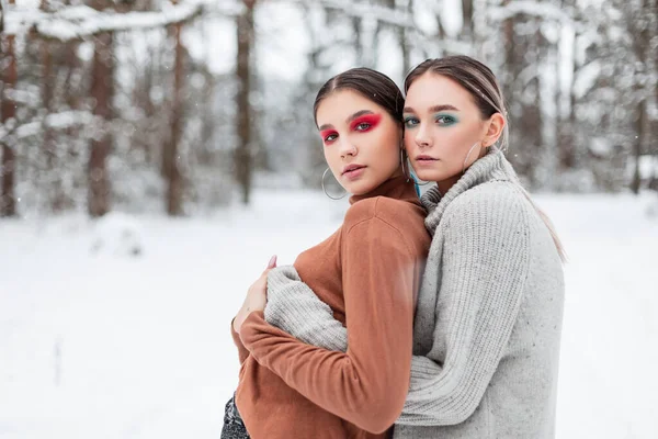 Eskimiş örgü bir süveterin içinde parlak makyajlı iki güzel genç kadın ormanda karla sarılıyorlar.