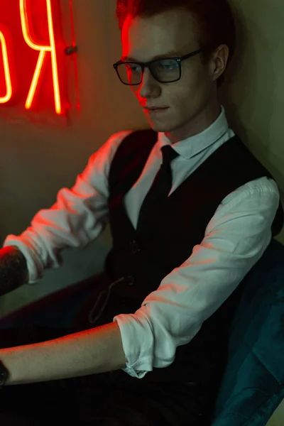 ファッショナブルなオフィスビジネス服の眼鏡を持つスタイリッシュなハンサムな若い男のモデルはネオン赤と緑の光と壁の近くの椅子に座っています — ストック写真