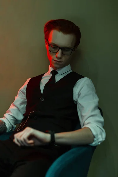 赤い髪のハンサムな若いビジネスマンの男でファッショナブルな服のスーツでシャツ ネクタイとベストは時計を見て 赤と緑の光で壁の近くの椅子に座って — ストック写真