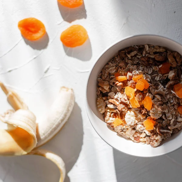 Lezzetli çiğ müsli, yulaf ezmesi, kurutulmuş kayısı, muz ve kuru meyveler beyaz kasede, üst manzara. Gün doğumunda vejetaryen yemeği.