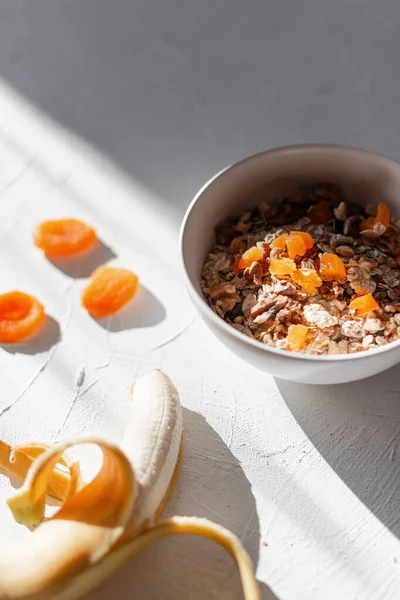 Lezzetli çiğ müsli, yulaf ezmesi, kurutulmuş kayısı, fındık ve muz sabah güneşiyle birlikte beyaz bir masada. Güneş doğarken sağlıklı yiyecekler. Günün başlangıcı ve Sağlık İçin Doğru Beslenme