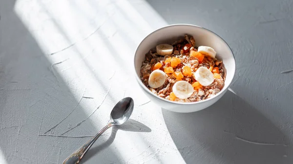 Lezzetli vejetaryen kahvaltı müsli, yulaf ezmesi, kurutulmuş kayısı, fındık ve muz beyaz bir kasede ve sabahın köründe beyaz bir masada bir kaşıkla. Sağlıklı çiğ gıda konsepti. Minimalizm