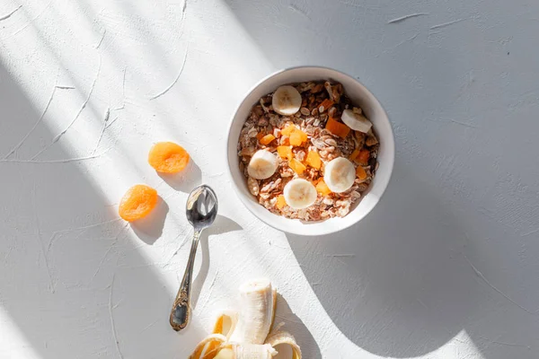 Kurutulmuş kayısı, muz ve kayıkla nefis sağlıklı kahvaltı müsli. Güneş ışığında beyaz bir masa ve sabahları gölgeler.