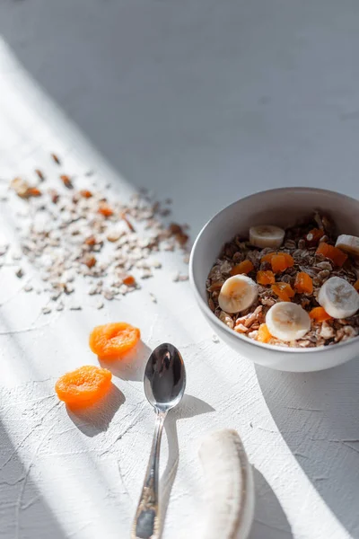 Yulaf ezmesi, vitamin, kurutulmuş kayısı, fındık ve muzlu müsli sağlıklı çiğ kahvaltı. Sabahın köründe beyaz bir masada kaşıkla. Vejetaryen yemeği