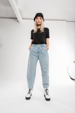 Kot pantolonlu, siyah tişörtlü, atölyede poz veren Panama şapkalı ve botlu güzel bir kadın manken. Yeni dış giyim koleksiyonu