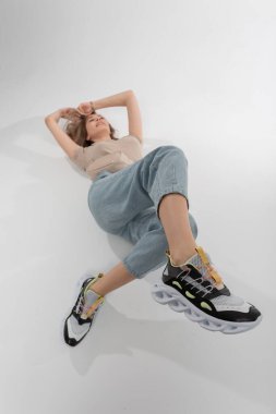 Spor ayakkabılarıyla şık yaz kıyafetleri giyen oldukça mutlu bir moda kadını beyaz bir arka planda yatıyor. Kot pantolonlu ve spor ayakkabılı güzel kadın.
